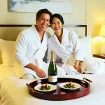 The image shows a man and a woman sitting on a bed in a hotel room. They are both wearing white bathrobes and are smiling at each other. On the bed, there is a wooden tray with two plates of food and a bottle of champagne. The tray is placed on the bedside table. The bed has white linens and there are two nightstands with lamps on either side of the bed. The walls are painted in a light color and there is an artwork hanging on the wall. The overall mood of the image is happy and relaxed.